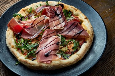 Jamon, roka, kırmızı sos ve gevrek kenarlı balzamik kaplı ahşap bir arka planda siyah bir tabakta servis edilen ahşap kaplı pizza. Bir çeşit İtalyan pizzası.