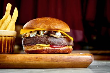 Lezzetli ve iştah açıcı burger, domates, marul, salatalık, eritilmiş peynir ve soğanlı Brioche ekmeği, ahşap bir tahtada patates kızartmasıyla servis edilir.