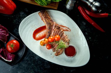Kemiğin üzerinde domuz filetosu, beyaz bir tabakta servis edilen kiraz domatesli ve kırmızı soslu. Koyu mermer arka plan. Barbekü et, restoran yemeği..