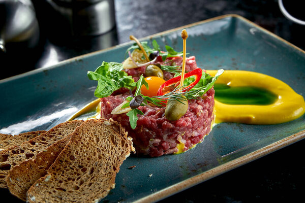 An appetizer before the main course - beef steak tartare served with croutons, cappers, yolk, pickles in a blue plate on a marble background. Restaurant food. Raw meat