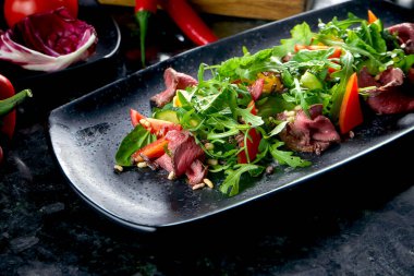 Biftek ve taze sebzeli sıcak salata, mermer arka planda siyah bir tabakta servis ediliyor. Restoran yemeği. Yaklaş, seçici odaklanma