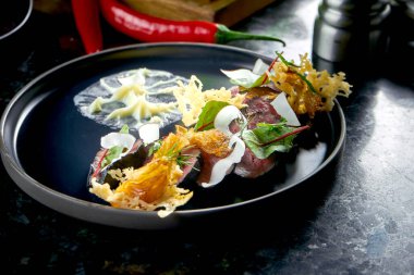 Dilimlenmiş siyah biftek, parmesan cipsi, hindistan cevizi ve beyaz sos, koyu mermer arka planda siyah bir tabakta servis ediliyor. Restoran yemekleri.