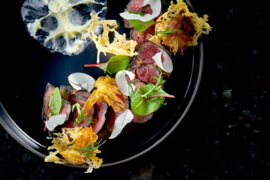 Dilimlenmiş siyah biftek, parmesan cipsi, hindistan cevizi ve beyaz sos, koyu mermer arka planda siyah bir tabakta servis ediliyor. Restoran yemekleri.