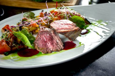 Beyaz tabakta servis edilen, sebze garnitürlü orta pişmiş sığır eti madalyonları. Koyu mermer arka plan. Barbekü et, restoran yemeği. Lezzetli biftek.