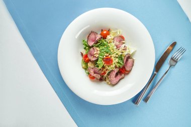 İştah açıcı, kızarmış biftekli sıcak salata, vişneli domates, parmesan ve çam fıstığı mavi masa örtüsünde beyaz bir tabakta.