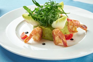 Avokado guacamole, karides, roka ve kiraz domatesli mavi masa örtüsü üzerinde beyaz bir tabak. Kapatın. Meksika yemeği