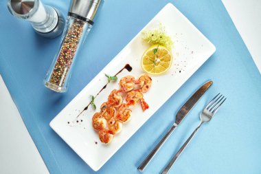 Mavi masa örtüsünde beyaz tabakta limonlu ızgara Akdeniz karidesi.