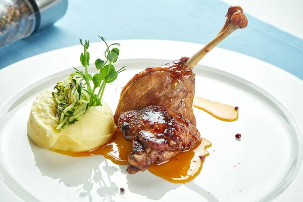 Duck leg confit with sweet sauce garnished with mashed potatoes in a white plate on a blue tablecloth