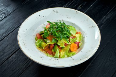 Karışık sebzelerden oluşan diyet ve vejetaryen salatası ve fıstık soslu marul, siyah ahşap arka planda beyaz bir tabakta servis ediliyor.