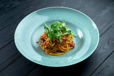 Geleneksel makarna, kıymalı makarna ve roka soslu domates, siyah ahşap arka planda mavi bir tabakta servis ediliyor. İtalyan mutfağı