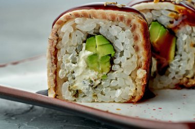 Lezzetli suşi rulo altın ejder krem peynir, avokado, yılan balığı ve unagi soslu gri arka planda beyaz bir tabakta. Japon yemeği. Sığ odak derinliği. Kapat.