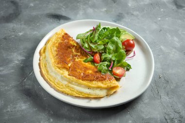 Klasik Fransız omleti, erimiş peynir ve salata karışımı, vişneli domates ve avokado, gri arka planda beyaz bir tabakta. Nefis kahvaltı.