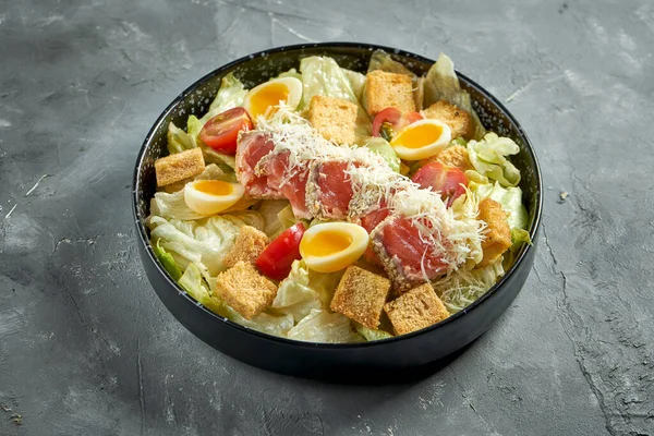 A classic American dish - Caesar salad with salmon, croutons, parmesan and tomatoes in a black plate on a gray background.
