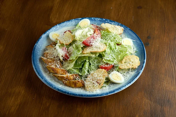 The classic American dish is Caesar salad with chicken, croutons, parmesan and tomatoes in a blue plate over a wooden background.