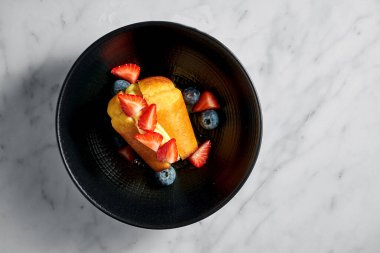 Kremalı Rum Baba keki, çilek dilimleri ve beyaz mermer bir masada siyah bir tabakta yabanmersini.