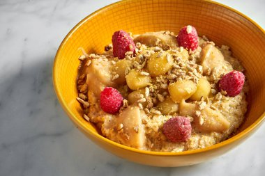 Lezzetli ve sağlıklı yulaf ezmesi, bal, elma, ahududu ve ay çekirdeği beyaz mermer arka planda parlak sarı bir kasede..