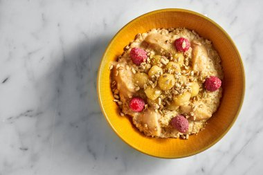 Lezzetli ve sağlıklı yulaf ezmesi, bal, elma, ahududu ve ay çekirdeği beyaz mermer arka planda parlak sarı bir kasede..