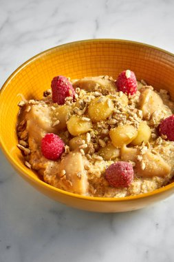 Lezzetli ve sağlıklı yulaf ezmesi, bal, elma, ahududu ve ay çekirdeği beyaz mermer arka planda parlak sarı bir kasede..