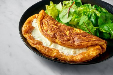 Beyaz mermer arka planda siyah tabakta salata karışımlı dolgun kremalı omlet.