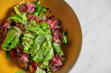 Orta pişmiş biftek dilimli taze salata, beyaz mermer bir masada sarı bir kasede kiraz domatesli.