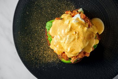 İştah açıcı ve sağlıklı kahvaltı. Avokadolu tost, fırında domates, haşlanmış yumurta ve hollandaise sosu mermer arka planda siyah bir tabakta servis ediliyor.