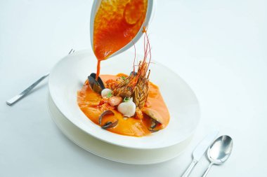İştah açıcı kırmızı Bouillabaisse çorbası beyaz bir masa örtüsü üzerinde beyaz bir tabakta servis edilen somon, istiridye, langoustine ve midye dolu bir kaseden dökülür. Deniz mahsullü domates çorbası.
