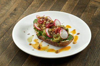 Avokado tostu, guacamole, turp, güneşte kurutulmuş domates ve çavdar ekmeğinin üzerinde tohumlar, ahşap arka planda beyaz bir tabakta servis ediliyor. Vejetaryen yemeği.