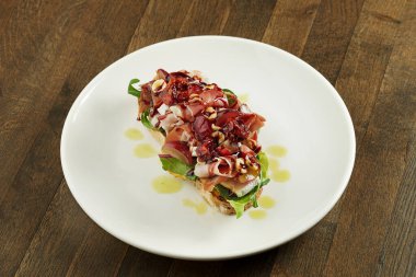 Jamon kabuklu, peynirli, karışık salatalı, üzümlü, güneşte kurutulmuş domatesli ve çavdar ekmeğinde fındıklı büyük sandviç, ahşap bir arka planda beyaz bir tabakta servis ediliyor. Nefis kahvaltı tostu