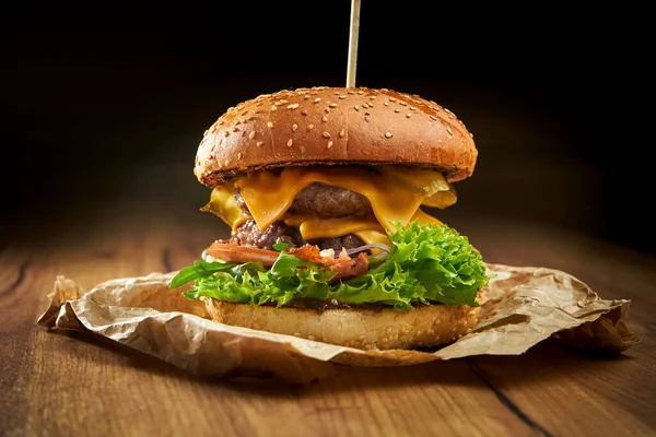 Juicy double burger with beef, salad, pickles, bacon and cheddar cheese, served on parchment on a wooden background. American fast food