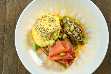 İştah açıcı kahvaltı, haşlanmış yumurta, somon ve ahşap arka planda beyaz bir kasede guacamole sosuyla süslenmiş Hollandaise soslu kızarmış ekmek.