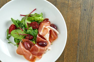 Jamon kabuğuyla karıştırılmış salata, zeytin ve kurutulmuş domatesler ahşap arka planda beyaz bir kasede..