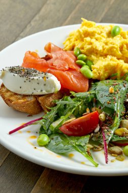 Lezzetli ve sağlıklı bir kahvaltı - çırpılmış yumurta ve krem peynirli tost ve somon, salatayı ahşap bir arka planda beyaz bir tabakta karıştırmak