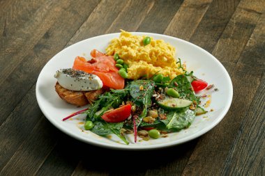 Lezzetli ve sağlıklı bir kahvaltı - çırpılmış yumurta ve krem peynirli tost ve somon, salatayı ahşap bir arka planda beyaz bir tabakta karıştırmak