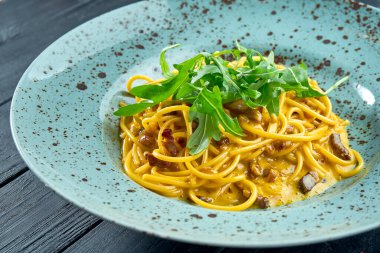 Pastırmalı İtalyan carbonara makarnası, çiğ yumurta sarısı, parmesan ve roka. Koyu arkaplanda mavi tabakta servis edilen ev yapımı makarna.