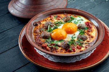 Klasik İsrail çırpılmış yumurtası - domatesli Shakshuka, otlar ve et kebabı, siyah arka planda kilden bir tabakta servis ediliyor..