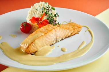 Sarı soslu kızarmış somon bifteği ve yanında pilav renkli arka planda beyaz bir tabakta..