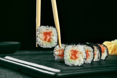 Maki suşisi ve somon. Klasik Japon mutfağı. Yemek servisi. Siyah arka plan. Çubuklar sushi tutar