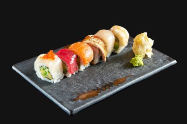 Farklı deniz ürünleri içeren gökkuşağı suşi rulosu: somon, yılan balığı, karides, istiridye, ton balığı ve avokado, seramik tabakta zencefil ve wasabi ile servis edilir. Siyah bir arka planda izole edilmiş. Japon yemeği.