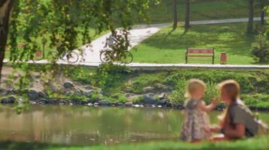 Genç bir çift gölün yanındaki parkta bisikletlerine biniyor. Ön planda, genç bir kadın çimlerin üzerinde küçük bir çocukla oynuyor. Güzel güneş parlıyor, yaz, rahatlama..