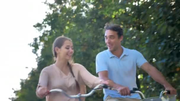 Casal jovem feliz em bicicletas em um contexto de árvores verdes. Um jovem e uma mulher estão sorrindo, se comunicando, rindo. Brilho de sol, romântico, amor. — Vídeo de Stock