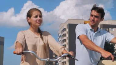 Bisikletli mutlu genç çift ileriye bakar, gülümser, güler. Kadın selamlaşmak için elini sallıyor. Yaz güneşli bir gün, bisiklet sürmek, yaşam tarzı.. 