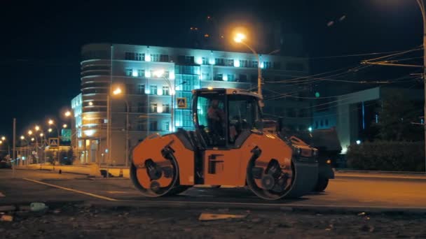 De roadroller staat tegen de achtergrond van nachtelijke stadsverlichting. Reparatie van wegdek, verbetering. Uitrusting voor wegenbouw. — Stockvideo