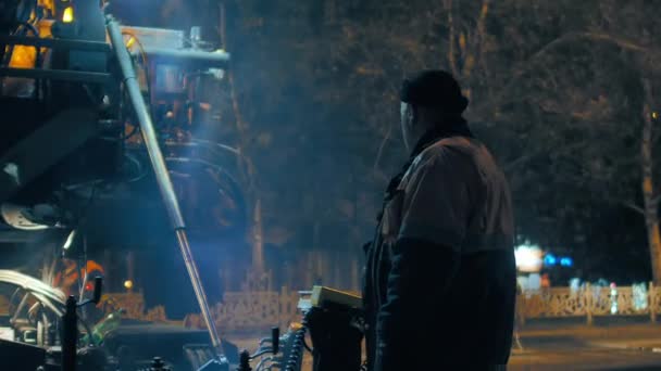 Wilayah Novosibirsk, 7 September 2019. Pekerja jalan di panel kontrol dari paver aspal. Sebuah mesin tumbuk bekerja di latar belakang. Uap dari aspal panas. Perbaikan jalan kota di malam hari. — Stok Video