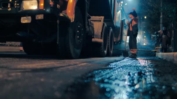 Région de Novossibirsk, 7 septembre 2019. Un travailleur de la route contrôle le processus de chargement du mélange d'asphalte dans un revêtement d'asphalte. Donne un ordre d'un geste de main. Réparation d'une route de ville la nuit. — Video