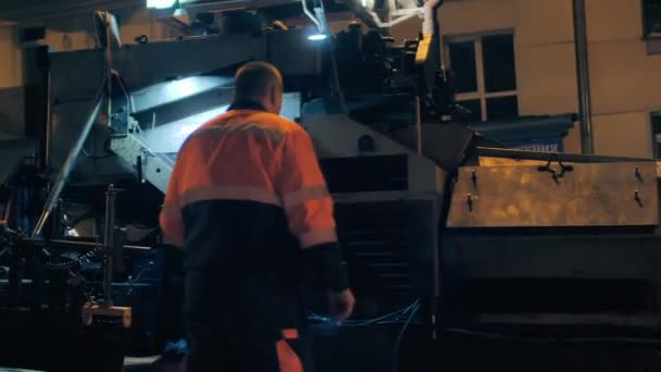 Vista trasera de un trabajador de la carretera frente a una pavimentadora de asfalto que pasa. Reparación de una carretera de la ciudad por la noche. Trabajos de construcción de carreteras. — Vídeos de Stock