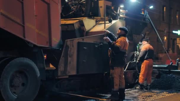 Novoszibirszk régió, 2019. szeptember 7. Egy útmunkás irányítja az aszfaltbeton keverék aszfaltbeton burkolatba töltésének folyamatát. Gőz forró aszfaltból. Városi út javítása éjszaka. — Stock videók