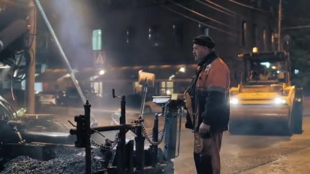 Novosibirsk, 7 de septiembre de 2019. Trabajador de carretera en el panel de control de pavimentos de asfalto. Reparación de carreteras en la ciudad por la noche. En el fondo, dos rodillos están colocando asfalto. — Vídeo de stock