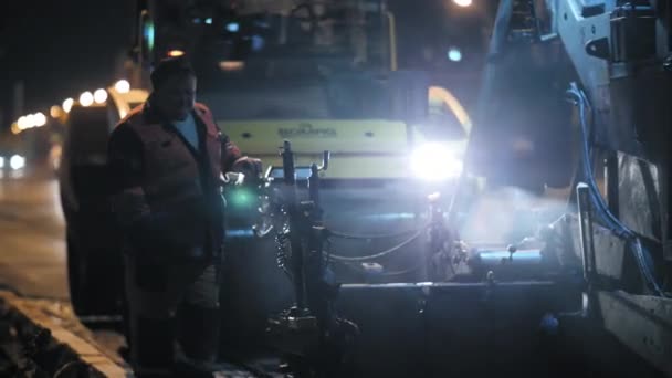 Novoszibirszk régió, 2019. szeptember 7. Útmunkás az aszfaltburkoló vezérlőpultnál. Egy úthenger dolgozik a háttérben. Egy egyenruhás férfi. Városi út javítása éjszaka. — Stock videók