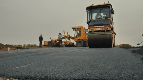 Novosibirsk bölgesi, 3 Eylül 2019. Yol silindiri asfalt seriyor. Arka planda bir not veren çalışıyor. Yol işçileri asfalt karışımı dağıtırlar. Bir yol silindiri davulunun yakın çekimi. Yol onarımı. — Stok video