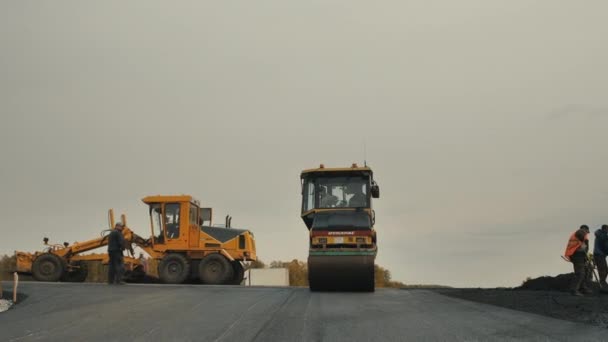 Gebiet Nowosibirsk, 3. September 2019. Die Straßenwalze verlegt Asphalt. Im Hintergrund arbeitet ein Grader. Straßenarbeiter verteilen Asphaltgemisch. Straßenreparatur, Straßenneubau. — Stockvideo